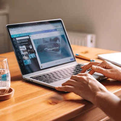 A person is working on the laptop. Next to the laptop is glass of water. Person is working on a light brown table, something with pictures on a canva website. A person is working on the laptop. Next to the laptop is glass of water. Person is working on a light brown table, something with pictures on a canva website.